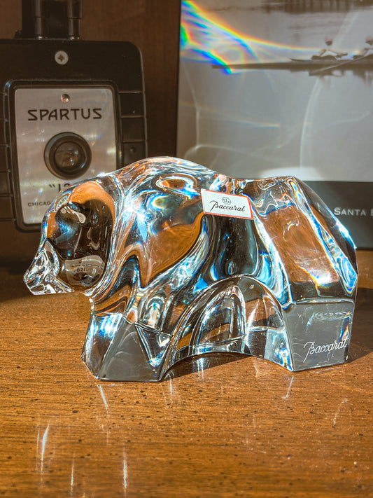 Crystal bear figurine on a wooden surface with camera and book in background. Baccarat Crystal Clear Oursons Wall Street Bear Paperweight Sculpture