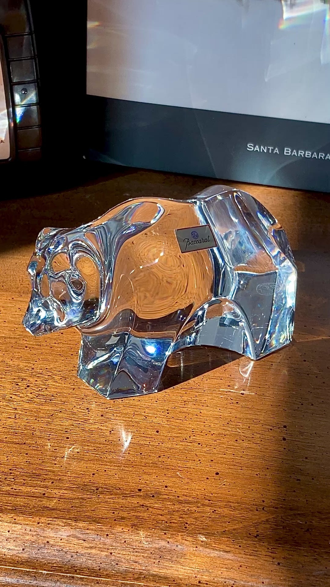 Crystal bear figurine on a wooden surface with camera and book in background. Baccarat Crystal Clear Oursons Wall Street Bear Paperweight Sculpture video of woman's hand turning the figurine and getting close ups.