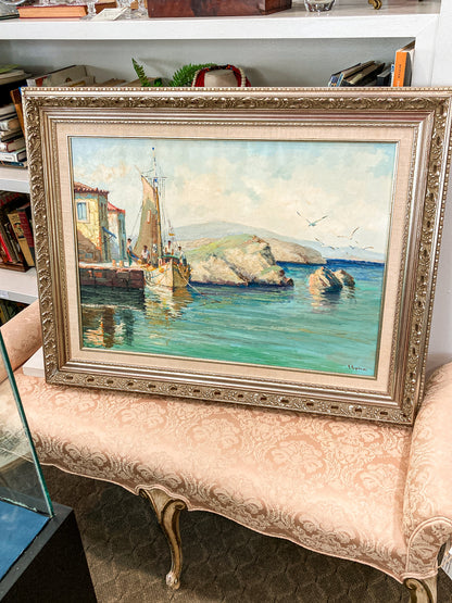 A Vintage 1960s Greece Village Seaside Boat Dock Landscape Signed Framed Oil Painting of a coastal harbor scene with a sailing boat, rocky island, and blue-green water, housed in an ornate gold-toned frame and displayed on a patterned pink bench in an antique shop.