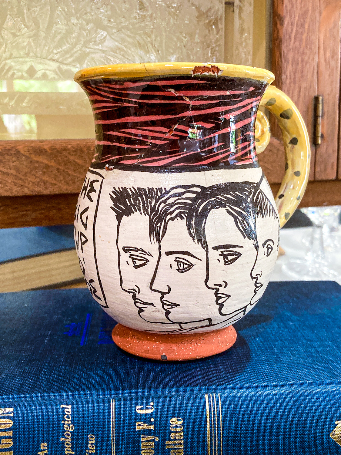 Ceramic mug with face designs on a blue book Vintage Daniel Postotnik Hand Potted Illustrative Portrait Mug As Found
