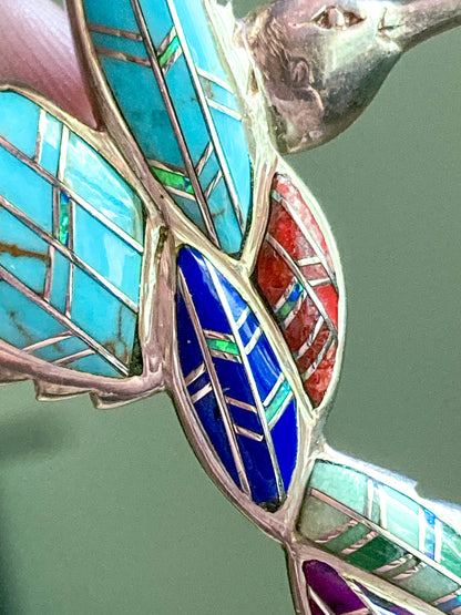 Close-up of a colorful Vintage Sterling Silver Turquoise Gemstone Mosaic Inlay Hummingbird Brooch brooch with turquoise, red, and blue enamel on a green background.