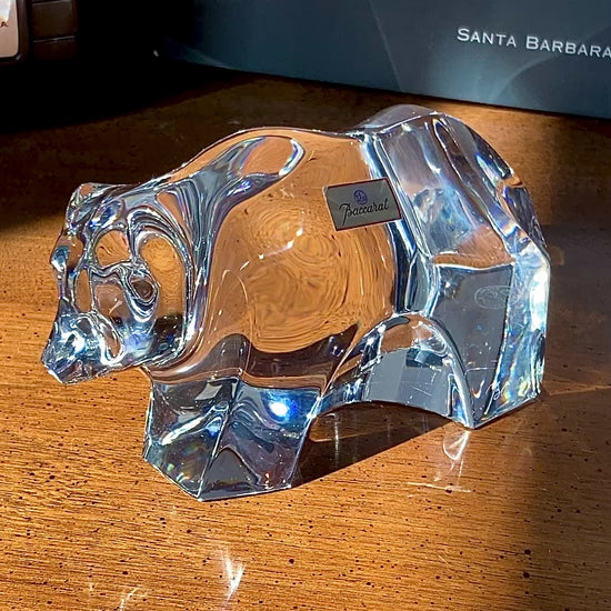 Crystal bear figurine on a wooden surface with camera and book in background. Baccarat Crystal Clear Oursons Wall Street Bear Paperweight Sculpture video of woman's hand turning the figurine and getting close ups.
